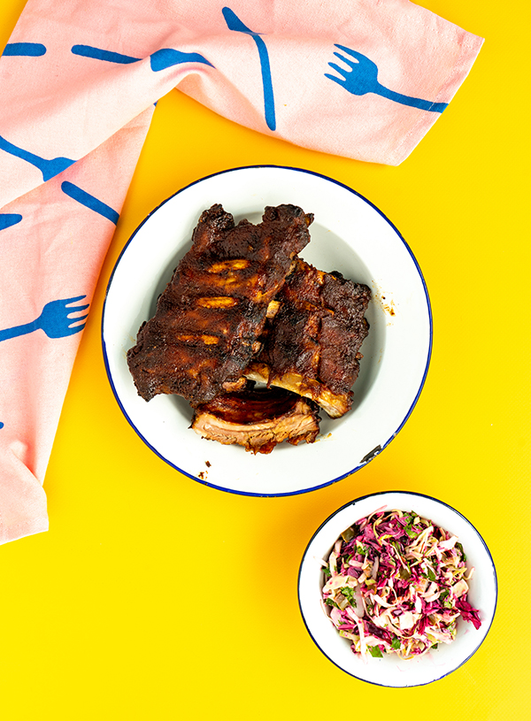 A plate of spareribs with a side of coleslaw and a pink tea towel draped to the side