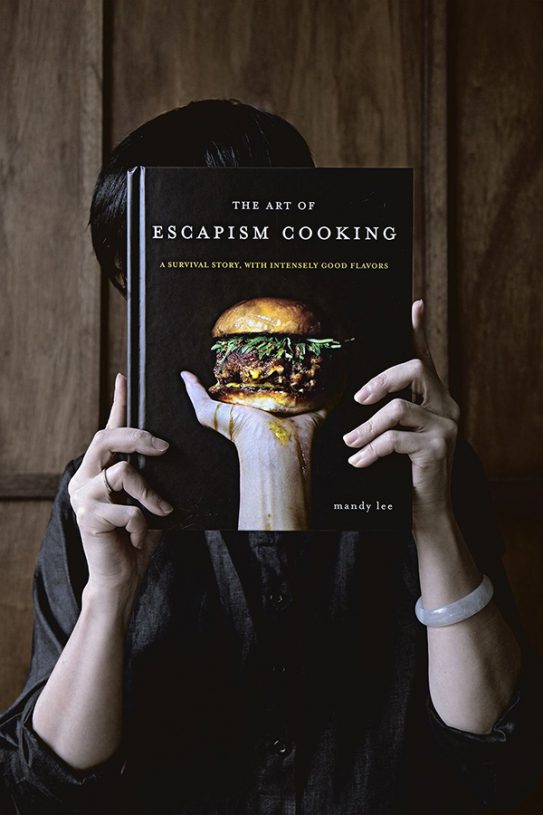 A picture of Mandy from Lady and Pups hiding behind a copy of her book The Art of Escapism Cooking