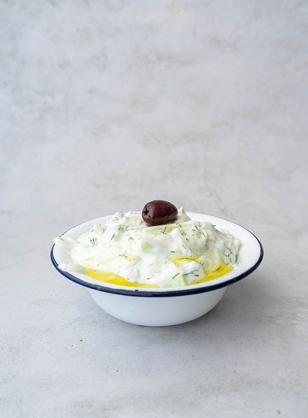 A white and blue rimmed enamel bowl filled with tzatziki and topped with a black olive 