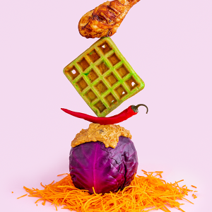 A fried chicken leg balanced on top of a green waffle, balanced on top of a chili pepper, balanced on top of a red cabbage covered in peanut sauce sat on top of shredded carrot on a pink background