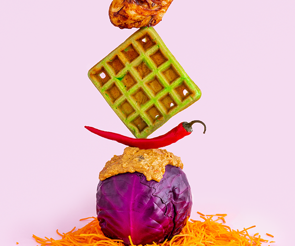 A fried chicken leg balanced on top of a green waffle, balanced on top of a chili pepper, balanced on top of a red cabbage covered in peanut sauce sat on top of shredded carrot on a pink background