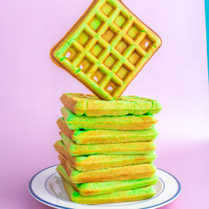 A white plate with a blue trim with a stack of green pandan waffles stacked on top, the top waffle appear to be floating on top of the stack vertically and then diagonally, there is a soft pink background with a slight edge of turquoise in the top right corner.