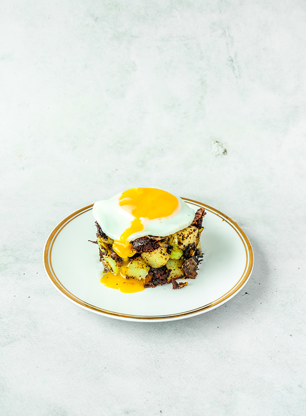A gold trimmed white plate on top of a cement background, on the plate is a little stack of potato, Brussel sprout and black pudding hash, topped with a fried egg. The yolk of the egg is drizzling down the front of the stack.