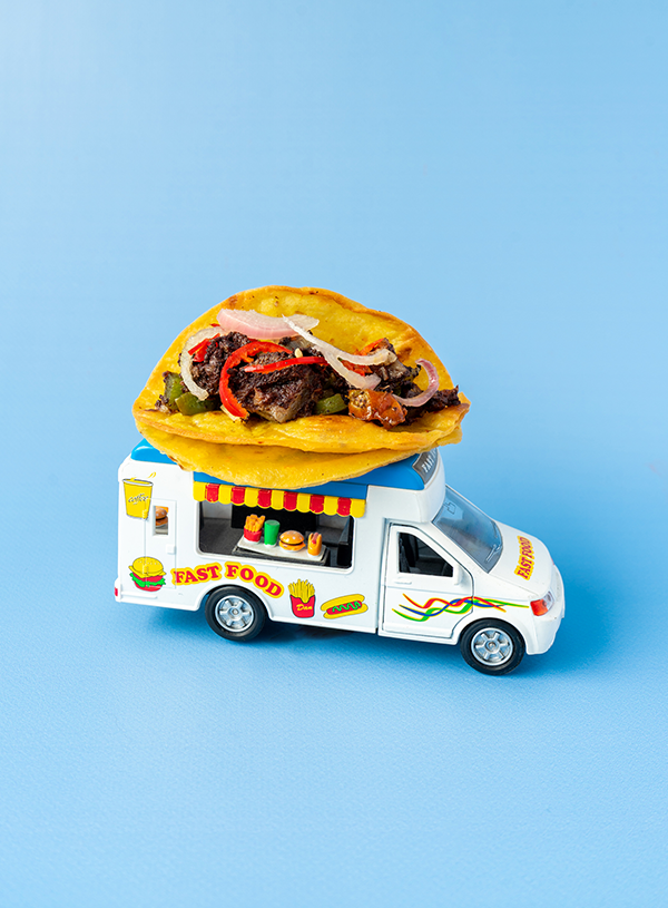 A brightly colored toy foodtruck with a taco filled with black pudding, green peppers, red chilies and pickled shallots atop a blue backdrop.