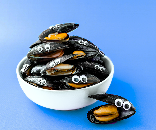 A stack of mussels in a white bowl on a blue background. The mussels have been cooked and are partially open, they have googly eyes stuck on their top shells.