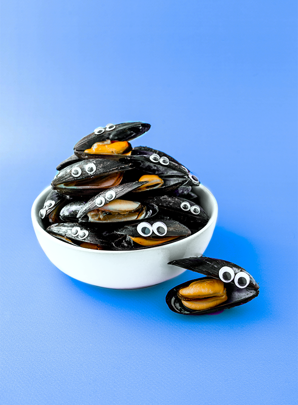 A stack of mussels in a white bowl on a blue background. The mussels have been cooked and are partially open, they have googly eyes stuck on their top shells.