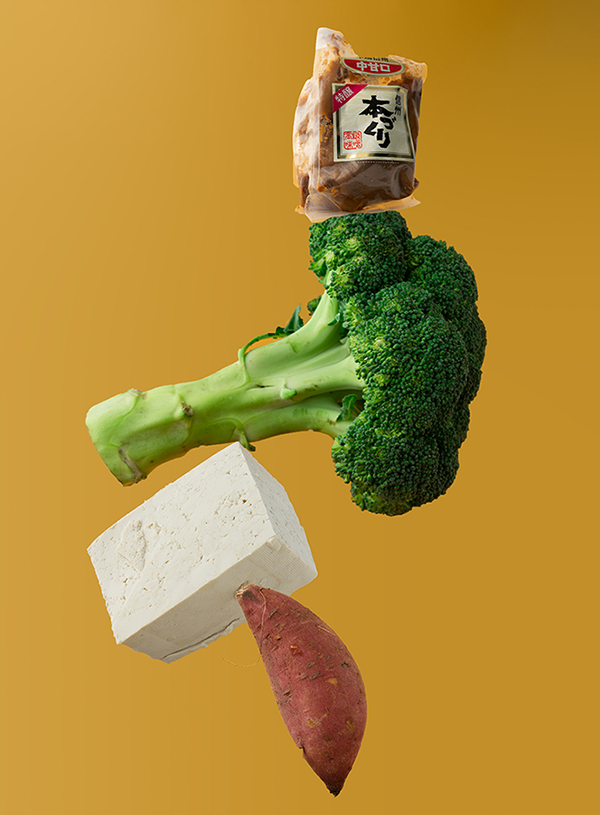 A sweet potato floating at the bottom om the image, with a block of tofu, brocolli and a packet of miso balancing on top on a mustard yellow background.