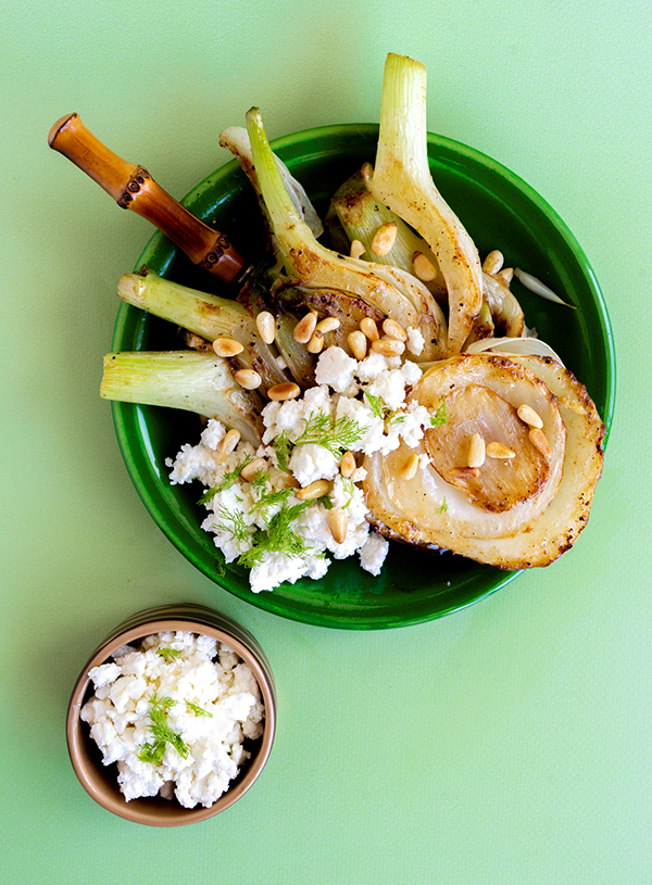 An overhead shot of a small green plate with caramelized fennel topped with roast pine nuts and crumbled feta and fennel fronds scattered across with a bamboo fork sticking out at the top, next to it is a small tan bowl filled with crumbled feta on top of a light green backdrop.