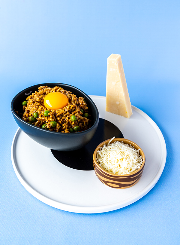 A blue background with a white plate with a black circle on top. On the plate to the right front there is a small brown and wood patterned dish with some grated Parmesan inside. To the back and left on the plate is an oval black bowl, filled with dark yellow brown rice dotted with peas, topped with a golden egg yolk. A wedge of parmesan has been set on it's backend to the right backend of the plate.