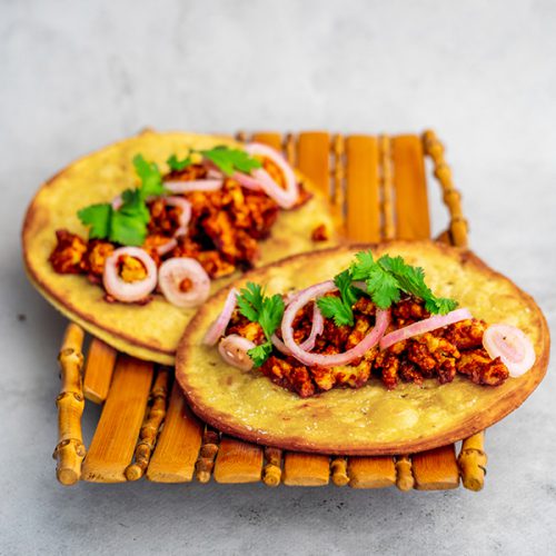 A bamboo platter on a cement backdrop, on the platter are two fried tacos with minced tofu and a red sauce as well as some pickled shallots and fresh coriander