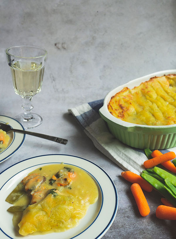 A retro 70s photography on a concrete background with a white plate with blue trim at the front with some fish pie on top. To the right of the plate are some carrots and sugar snaps. In the background is a glass of wine and to the right side there is an oven dish of fish pie set atop a white and blue dish towel.