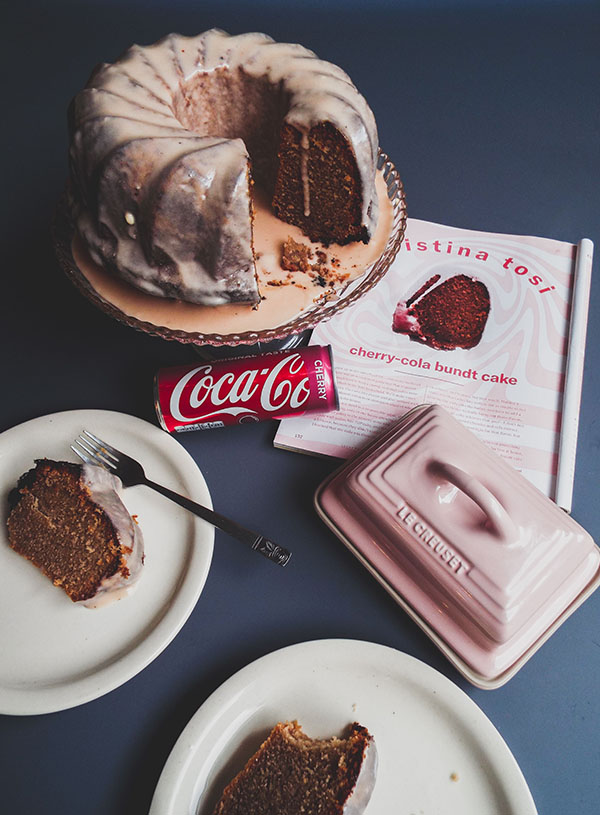 A cherry cola Bundt cake surrounded by slices of cake, a magazine, a pink butter dish and an empty can of cherry cola