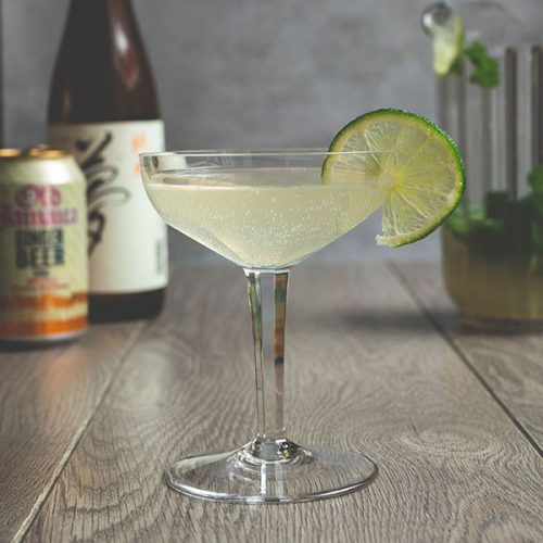 A champagne glass filled with the Japanese mule cocktail with a thin slice of lime on the rim, with a bottle of sake and a can of ginger beer in the background to the left and a jug of Japanse mule cocktail in the background to the right