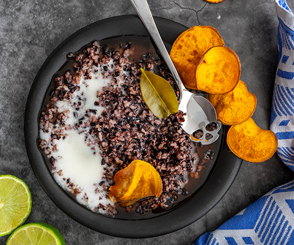 A black plate with black rice pudding and white coconut milk with a skull shaped teaspoon and a lime leaf, with fried sweet potato chips and a halved lime on the side