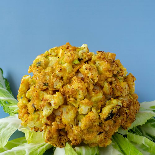 A head of cauliflower covered in cauliflower fritters, surrounded by its own leaves, on a blue background.