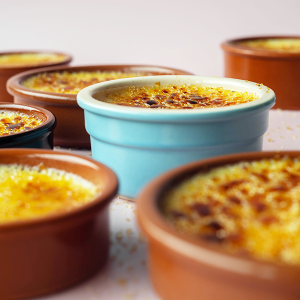 Seven bowls of crème brûlée sit together on a pink backdrop. The bowls are different heights. All the brown bowls are out of focus. One blue bowl in the center of the image is in focus.