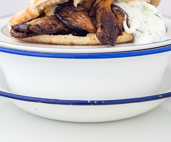 A close up of five white bowls and plates with blue trim, topped with a pita bread cut through the middle filled with roasted oyster mushrooms and tzatziki.