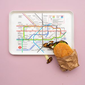 A server tray with the London tube map on it and a paper wrapper jerk bun perched on the bottom right side of it with mushrooms and mayonaise spilling out around it.