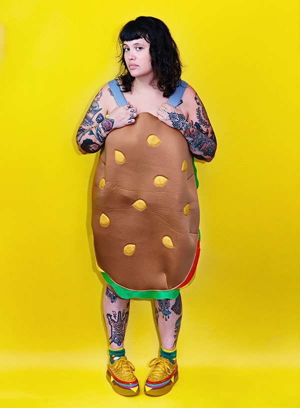 Dorothy Porker, a tattooed brunette with bangs, wearing nothing but a hamburger costume and hamburger sneakers on a bright yellow background.