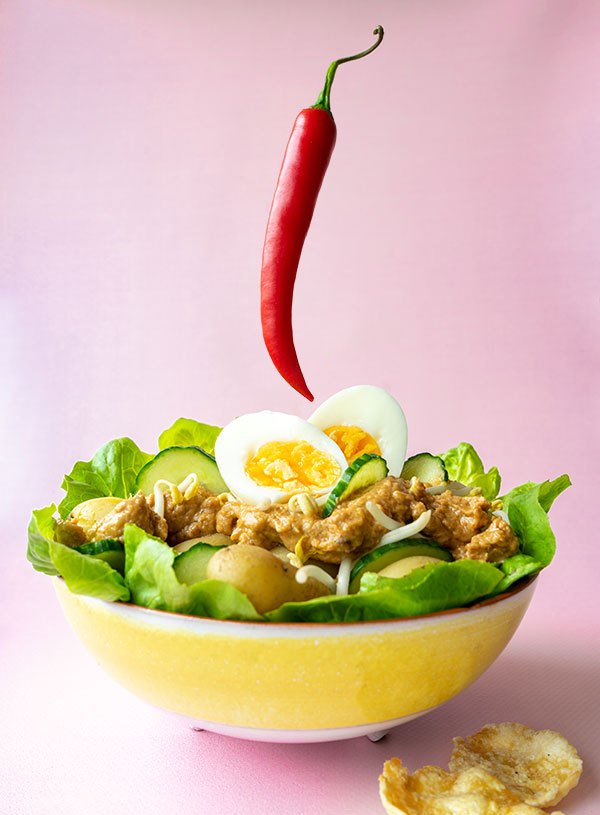 A yellow ceramic bowl with white trim filled with gado gado (lettuce, cucumber, new potatoes, bean sprouts, a soft boiled egg and a peanut-based sauce) with a red chili floating above it on a soft pink backdrop.