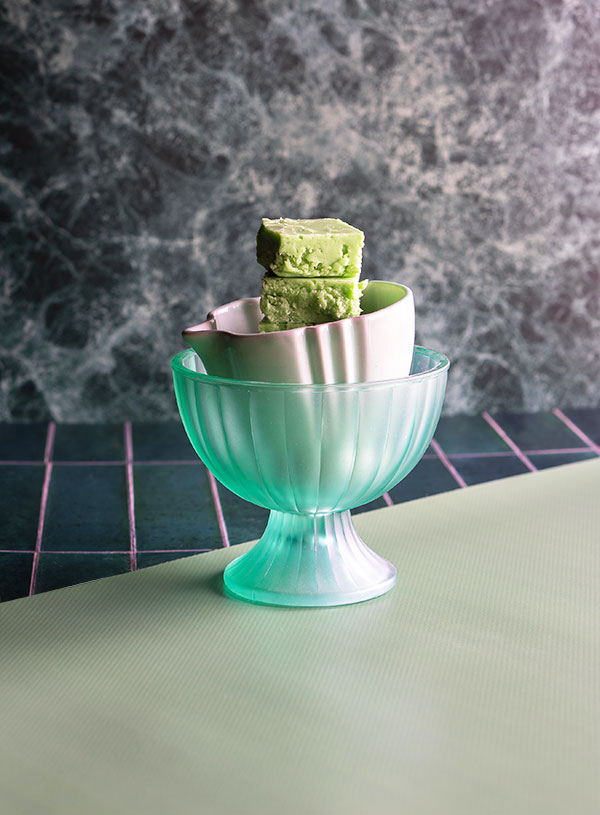 A translucent bright turquoise ice cream bowl, with a soft mint green ceramic sauce bowl inside of it, which has a small stack of green pandan fudge inside of it on a green marble, tile and mossy green background.