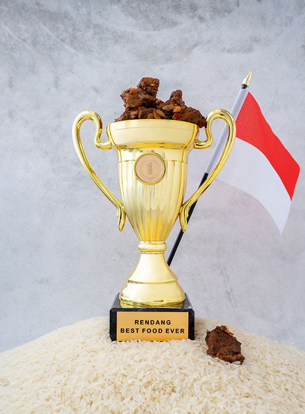 A golden number 1 trophy that says "Rendang - Best Food Ever" on top of a mountain of rice. The trophy is filled with rendang and is flanked by an Indonesian flag while there is a marble backdrop.