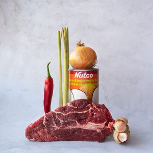 A blade of pot roast beef with a fresh root of galengal posed next to it and a red chili pepper and lemon grass stems posed behind it, alongside a can of Nutco coconutmilk topped by a sweet onion on a marble backdrop.