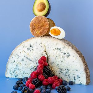 Half a wheel of blue cheese with a crack in the middle that has red fruits flowing out of it, topped with a round of crispy tofu and half a hard boiled egg and an avocado balancing on top on a blue backdrop.
