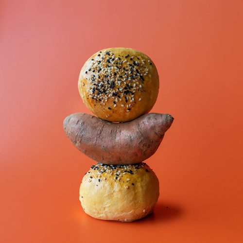 A stack of two sweet potato buns topped with everything bagel mix and a sweet potato on a bright orange background.