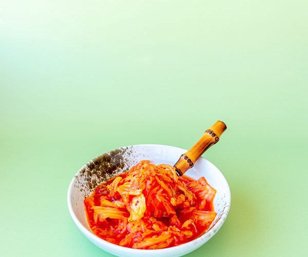 A small ceramic bowl, white with some splashes of brown, with some very bright kimchi and a bamboo handled fork sitting inside of it, on a mint green backdrop.
